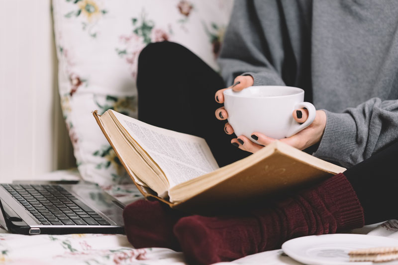 woman reading book that's placed on feet, while holding cup of coffee 
