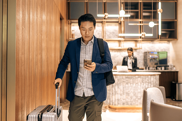 Man in an airport lounge looking at his cellphone while holding his rolling luggage.