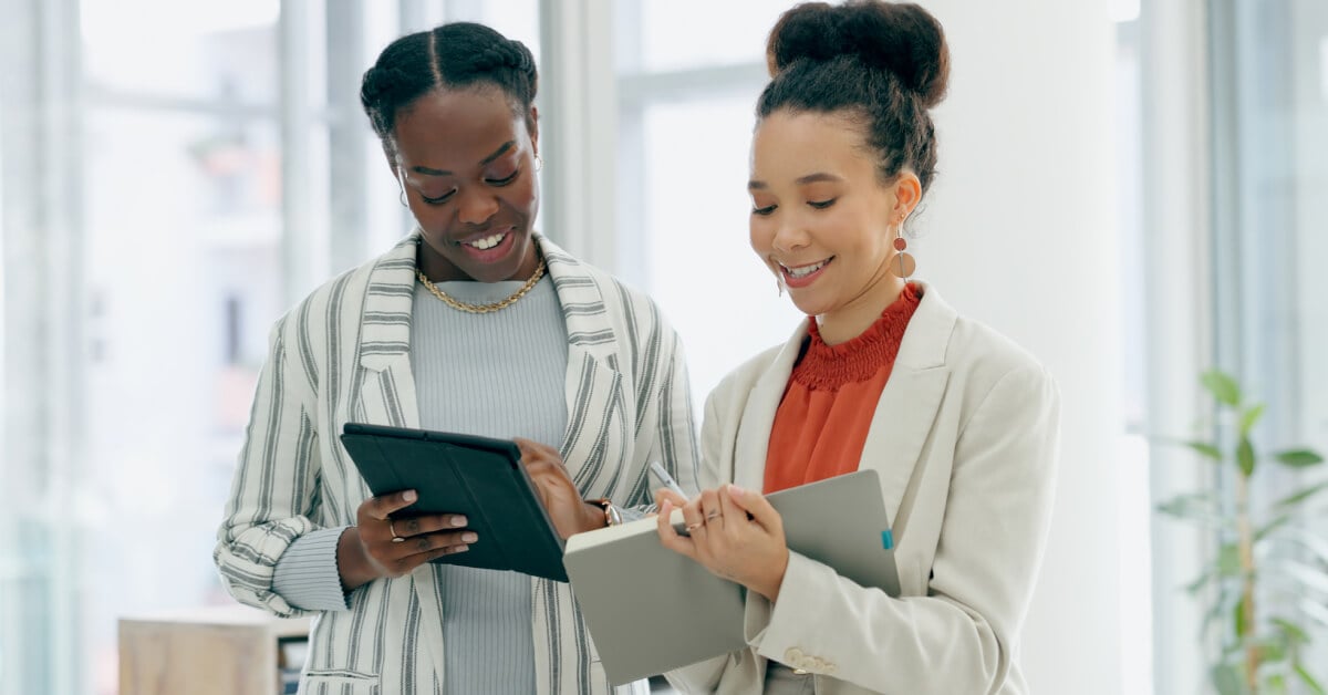 Two smiling women standing in a brightly lit office interacting with tablet computers.