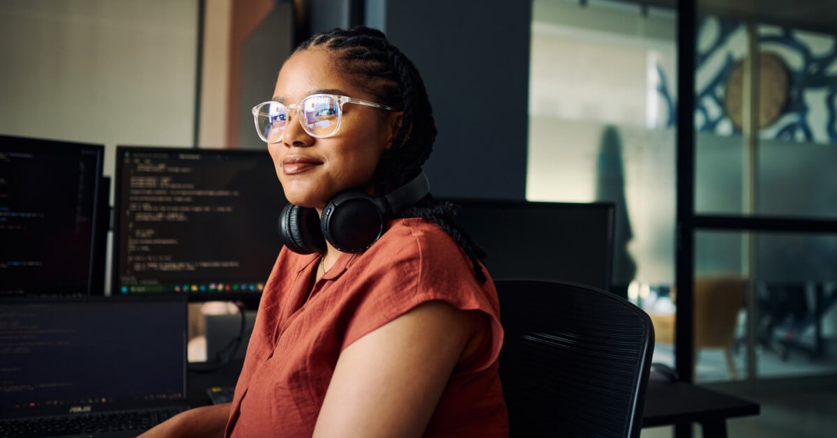 Person with braided hair and headphones around their neck sits at a desk in a modern office, surrounded by multiple computer monitors displaying lines of code. The person wears a loose-fitting rust-or