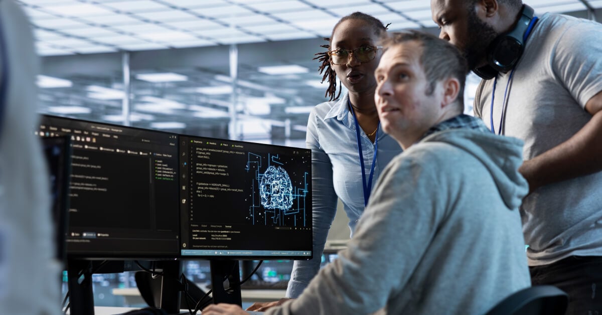 A group of office workers engaged in a heated discussion in front of a set of monitors.