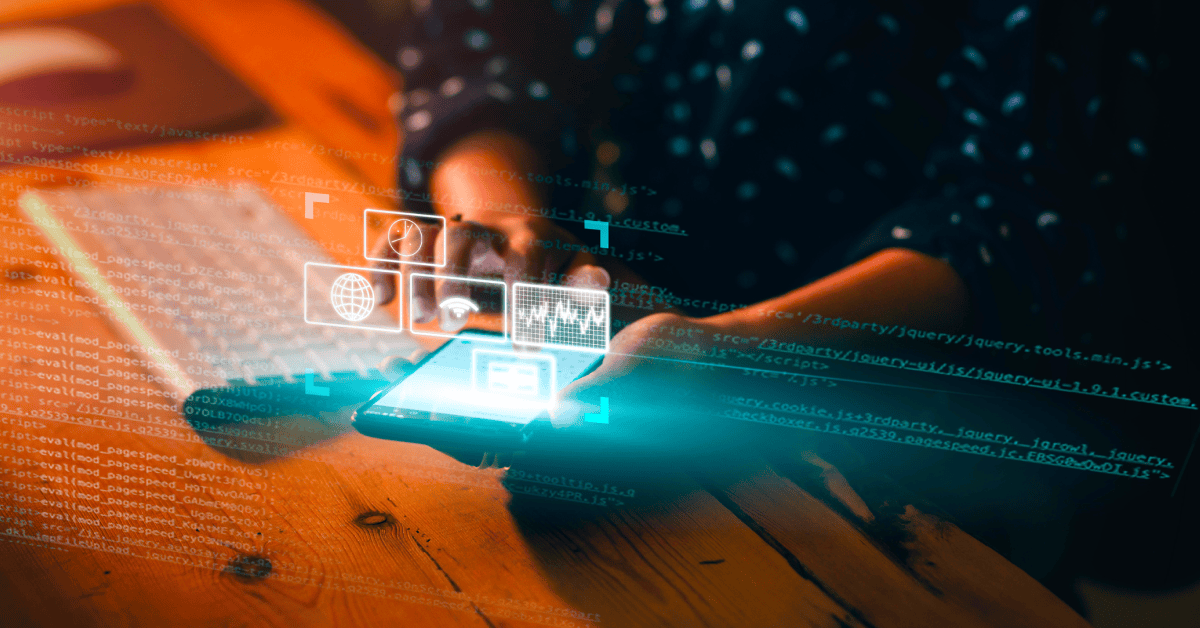 Hands using a smartphone on a wooden desk with a keyboard in the background; digital holographic icons for analytics, globe, and files appear above the phone alongside overlaid code snippets