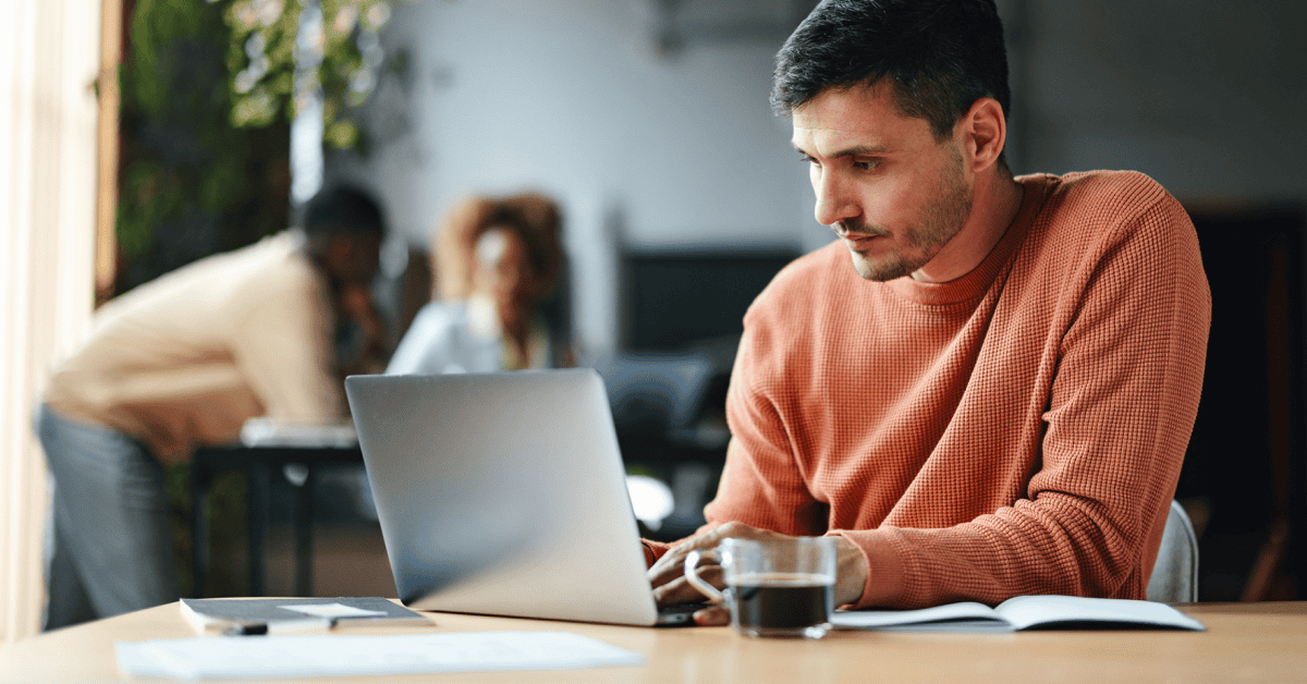 Person in a rust-colored sweater working on a laptop at a table with a notebook, smartphone, pen, and a glass mug of coffee; two people are conversing in the background at another desk.