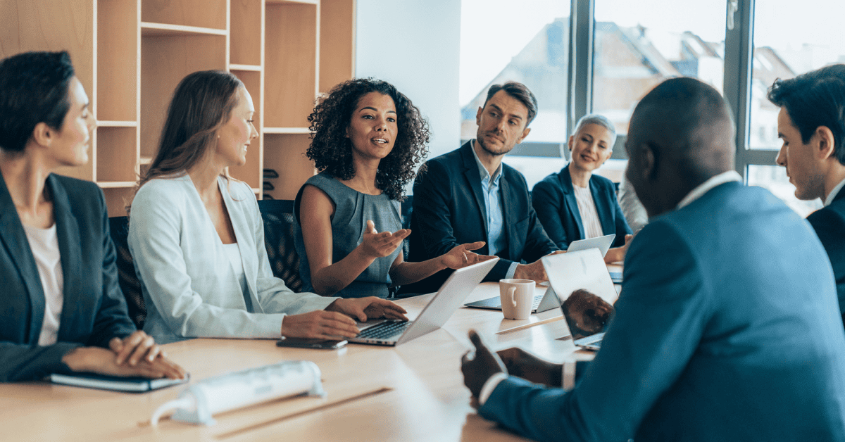 Group of professionals in business attire seated around a conference table with laptops, notebooks, and coffee cups, engaged in discussion in a modern office with large windows and shelves in the back