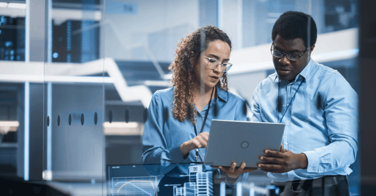 A man and woman standing in a modern office environment, collaborating and looking at a laptop together. There are large glass walls, reflections and digital interfaces visible.