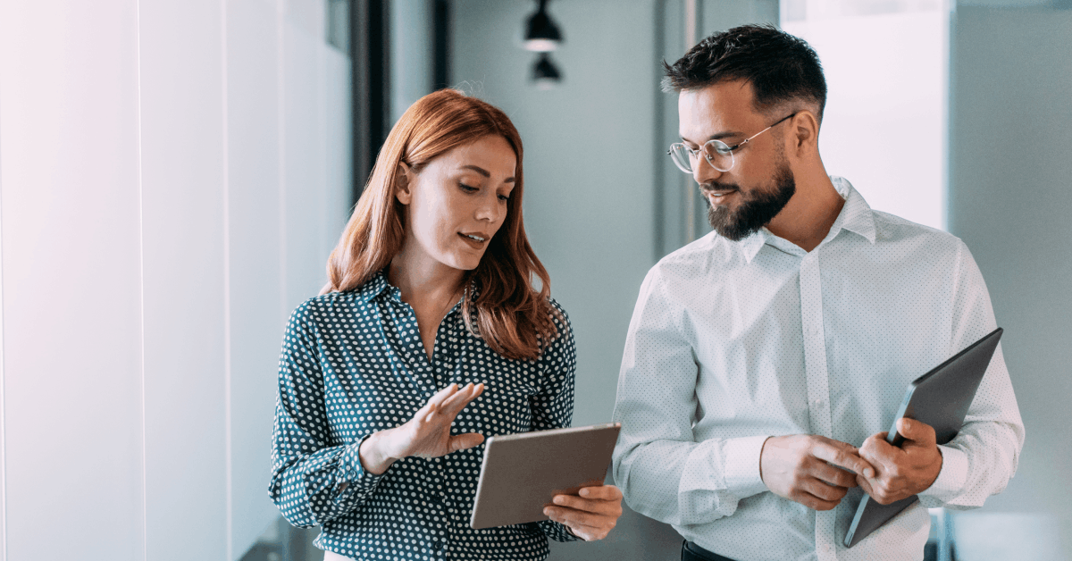 Two colleagues standing in a modern office hallway, engaged in conversation. One person is holding a tablet while gesturing with their hand, and the other person is holding a closed laptop.