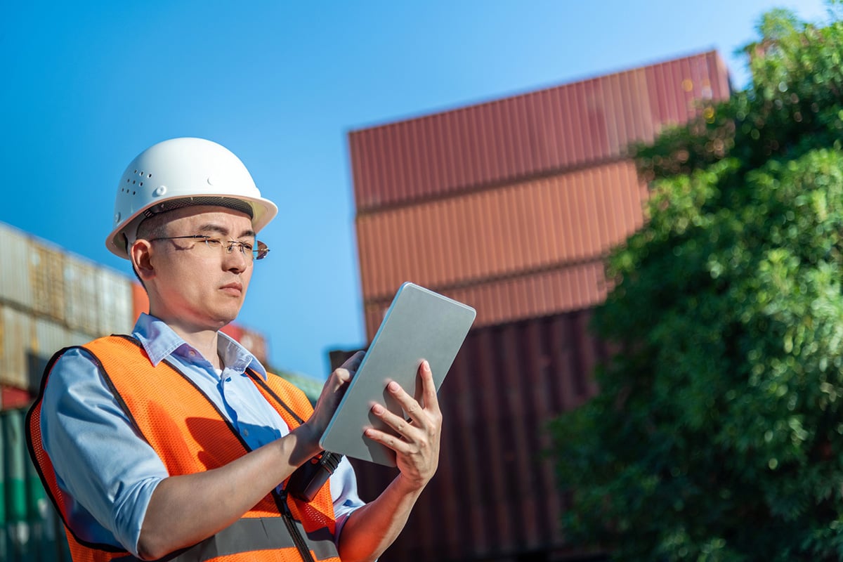 A person wearing a white safety helmet and an orange reflective vest is standing outdoors at a shipping yard, holding and using a tablet device. Shipping containers are stacked in the background.