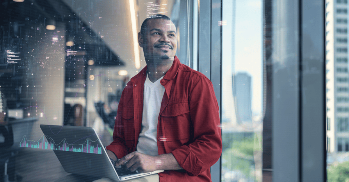 A person wearing a red jacket sits indoors by a large window, working on a laptop. The background shows a modern office setting with city buildings visible through the glass.