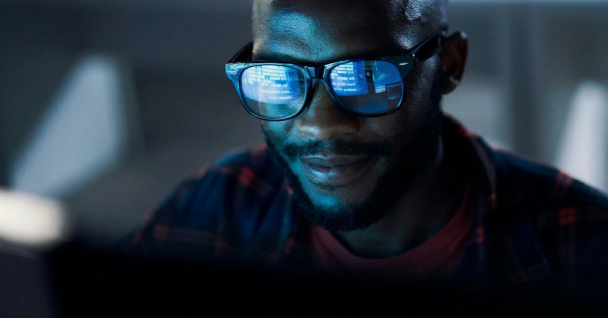 Gros plan sur une personne portant des lunettes, dont les verres reflètent des lignes de code, travaillant sur un ordinateur dans un environnement faiblement éclairé.