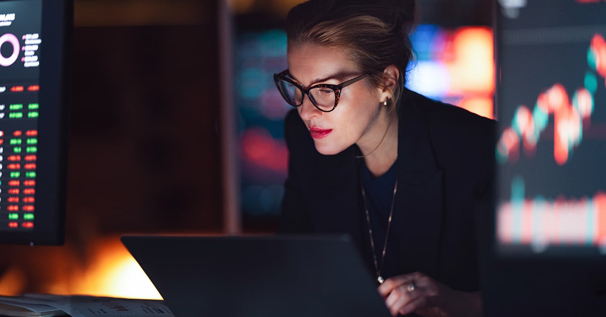 A woman wearing a business suit in a darkened room is typing into a laptop.