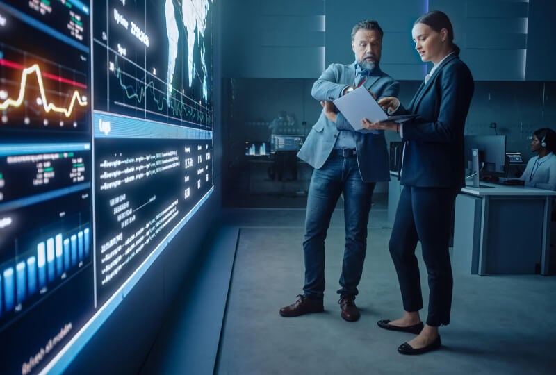 Man and woman interacting with a laptop held by the woman while standing in front of a wall of monitors.