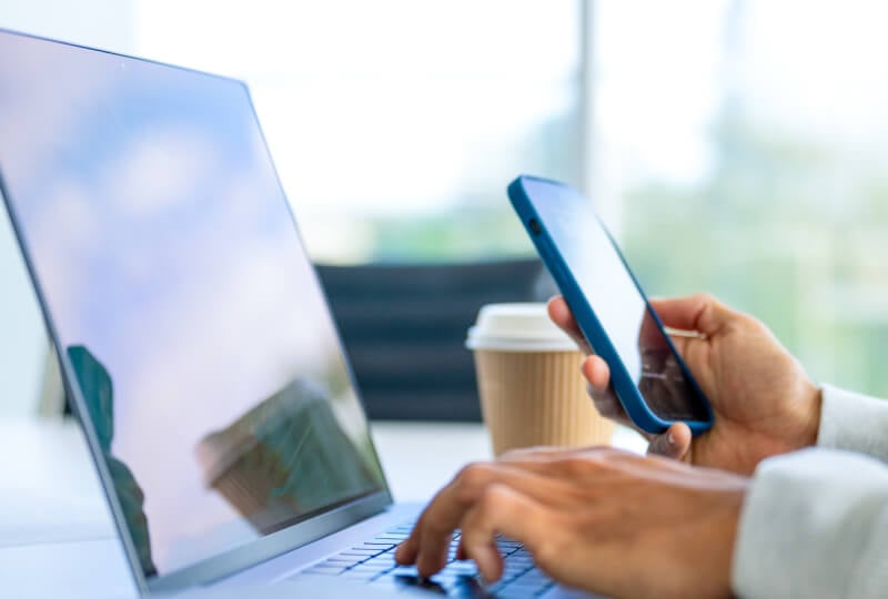 Closeup of a person's hands interacting with a laptop keyboard and smart device.