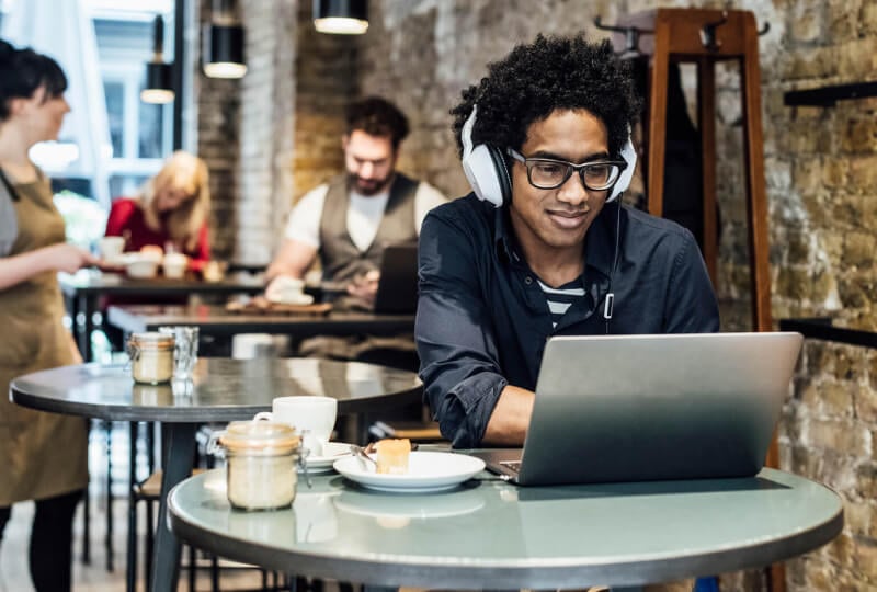 Smiling man wearing headphones and working on a laptop in a coffee shop.
