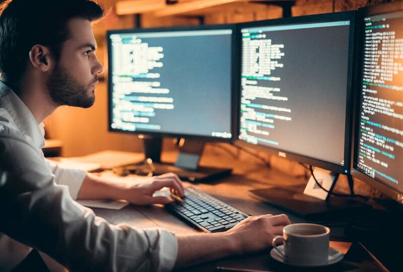 Man sitting in a dimly lit room working at a multi monitor computer.