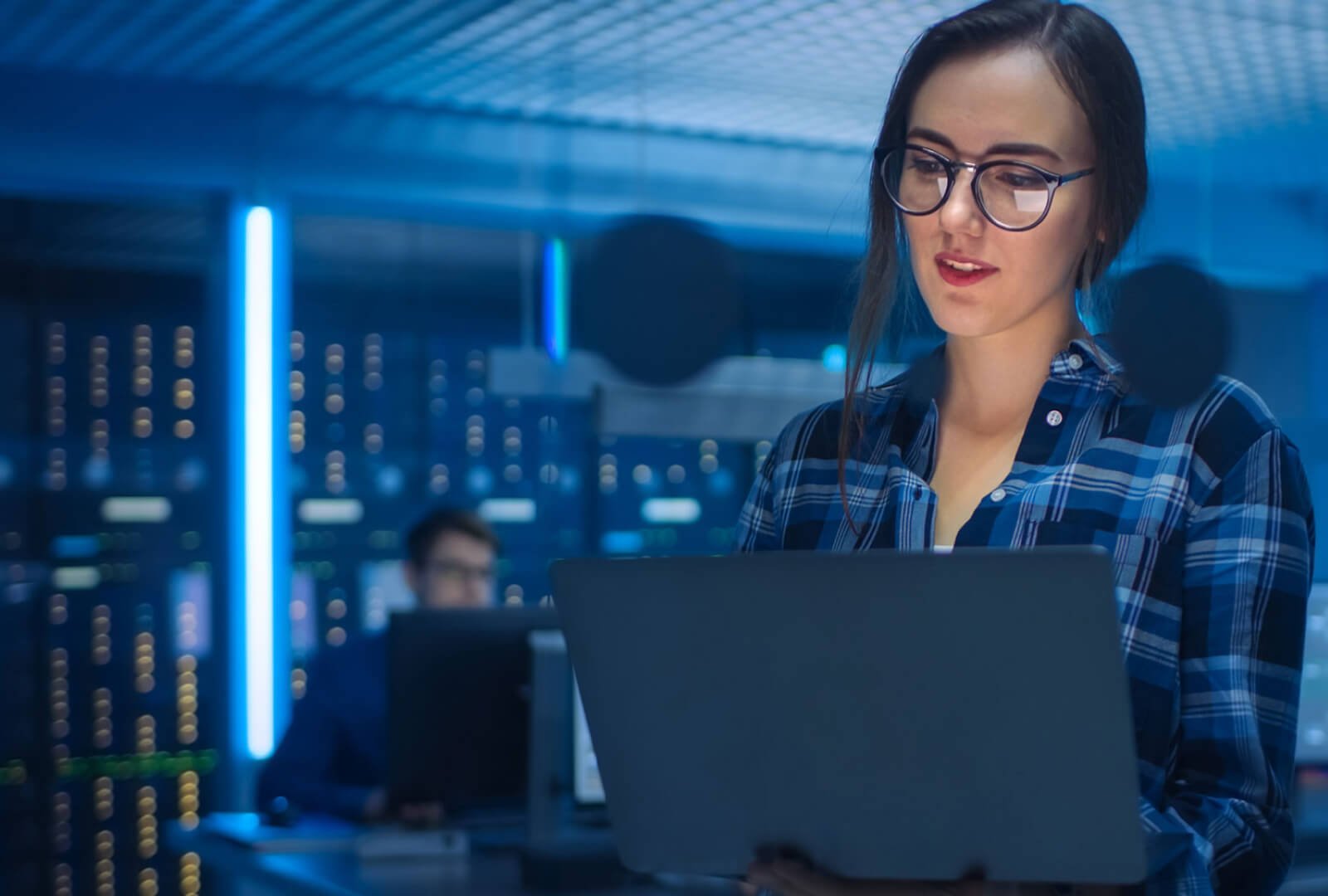 Woman wearing glasses in a server room interacting with a laptop.