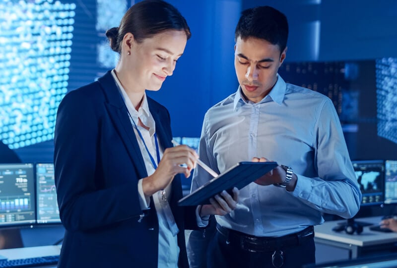 Man and woman in a server room interacting with a tablet computer held by the woman.