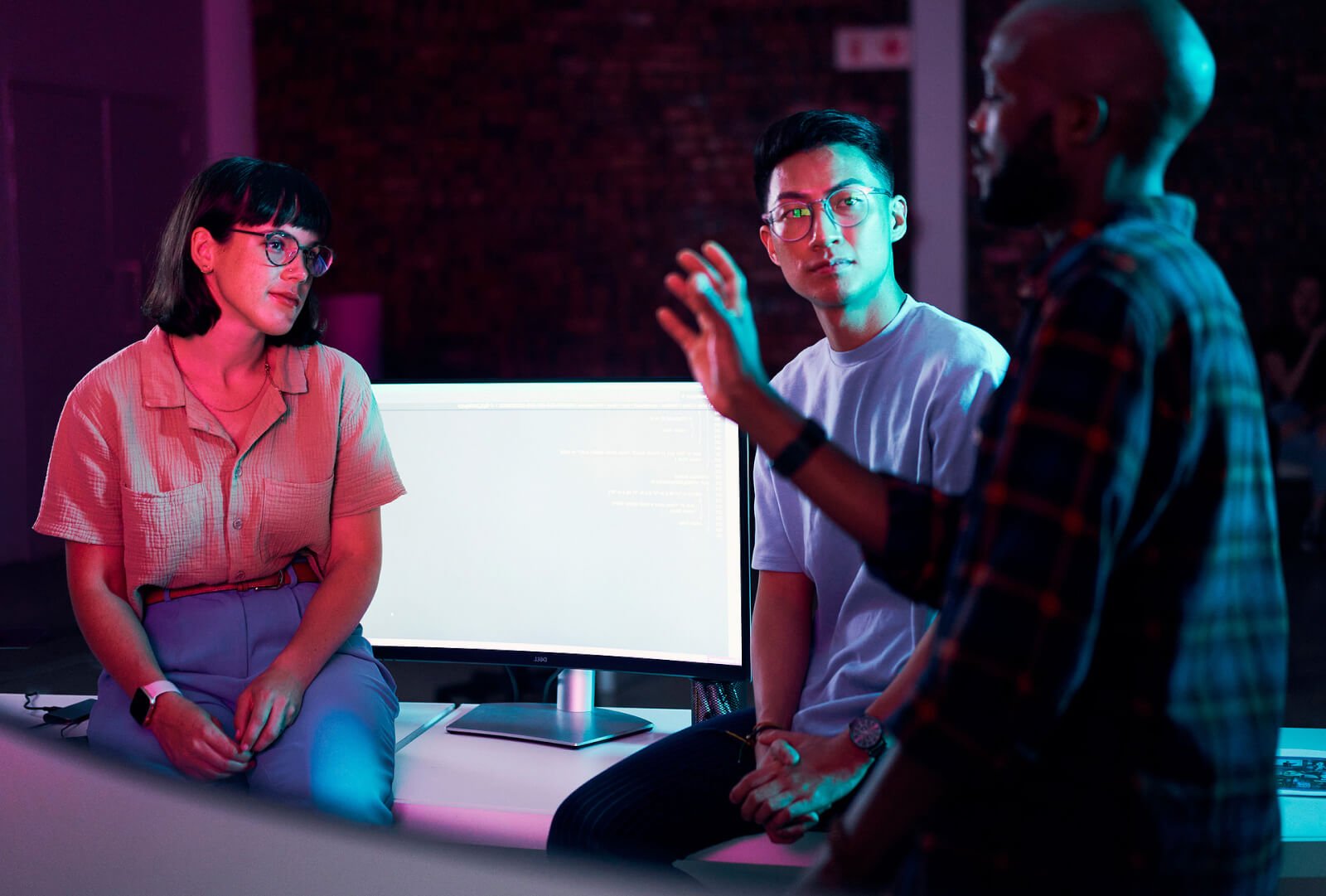 Three people sitting at a computer desk discussing something.