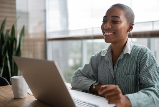 Smiling woman sitting in a brightly lit office and looking towards a laptop screen.