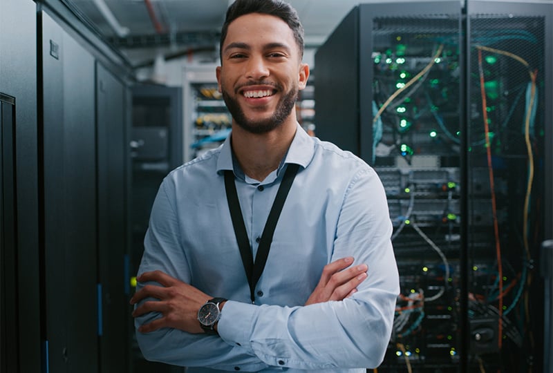 A man standing with arms crossed in a server room, wearing a light blue dress shirt and a lanyard. Behind them are server racks with visible cables and glowing indicator lights.
