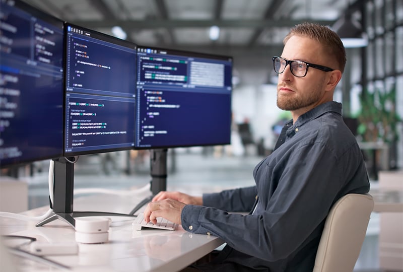 A man sitting at a desk in a modern office, working on a computer with three large monitors displaying lines of code. The workspace is bright and open, with plants visible in the background.