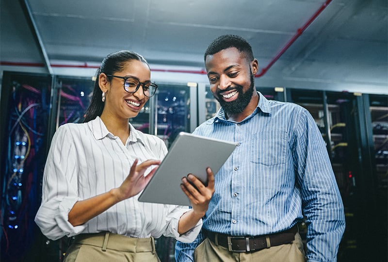 Two people standing in a server room, both wearing business attire. One is holding and using a tablet, while the other is observing. Server racks with visible cables are in the background.