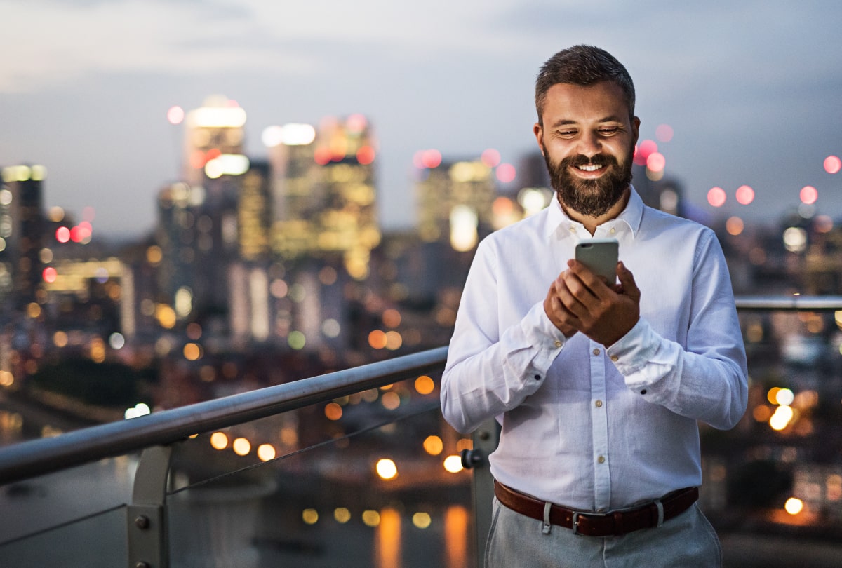 Smiling man standing on a rooftop patio interacting with his smartphone.
