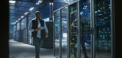 A man walking through a modern data center, holding a tablet. They are surrounded by tall glass server racks with glowing indicator lights. The environment is spacious and well-lit.