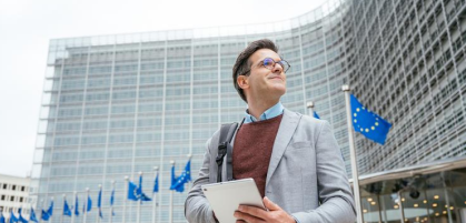 A man standing outside a modern office building holding a tablet and wearing a backpack. Multiple European Union flags are visible, indicating an EU-related location.