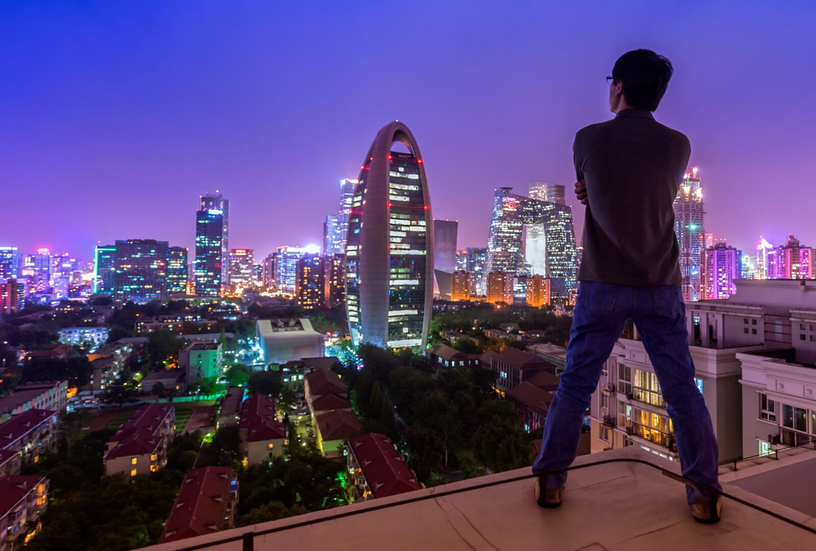 Man standing on a rooftop overlooking a cityscape at night.