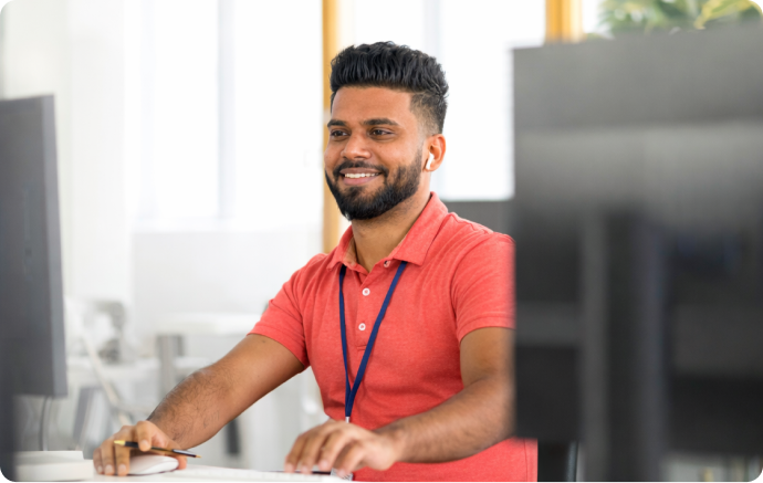 A person wearing a red polo shirt and a lanyard is sitting at a desk, working on a computer in a modern office environment. The background is bright, with natural light coming through the windows. Computer monitors are visible in the foreground and background.