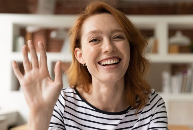 A smiling woman sitting indoors, wearing a black and white striped shirt, and raising her hand as if waving or greeting someone. Shelves with various items are visible in the background.