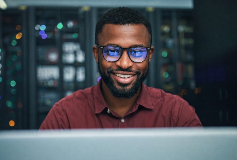 A man sitting in front of a computer in a server room or data center. He is wearing a maroon button-up shirt, and behind him are racks of servers with various lights and equipment visible.
