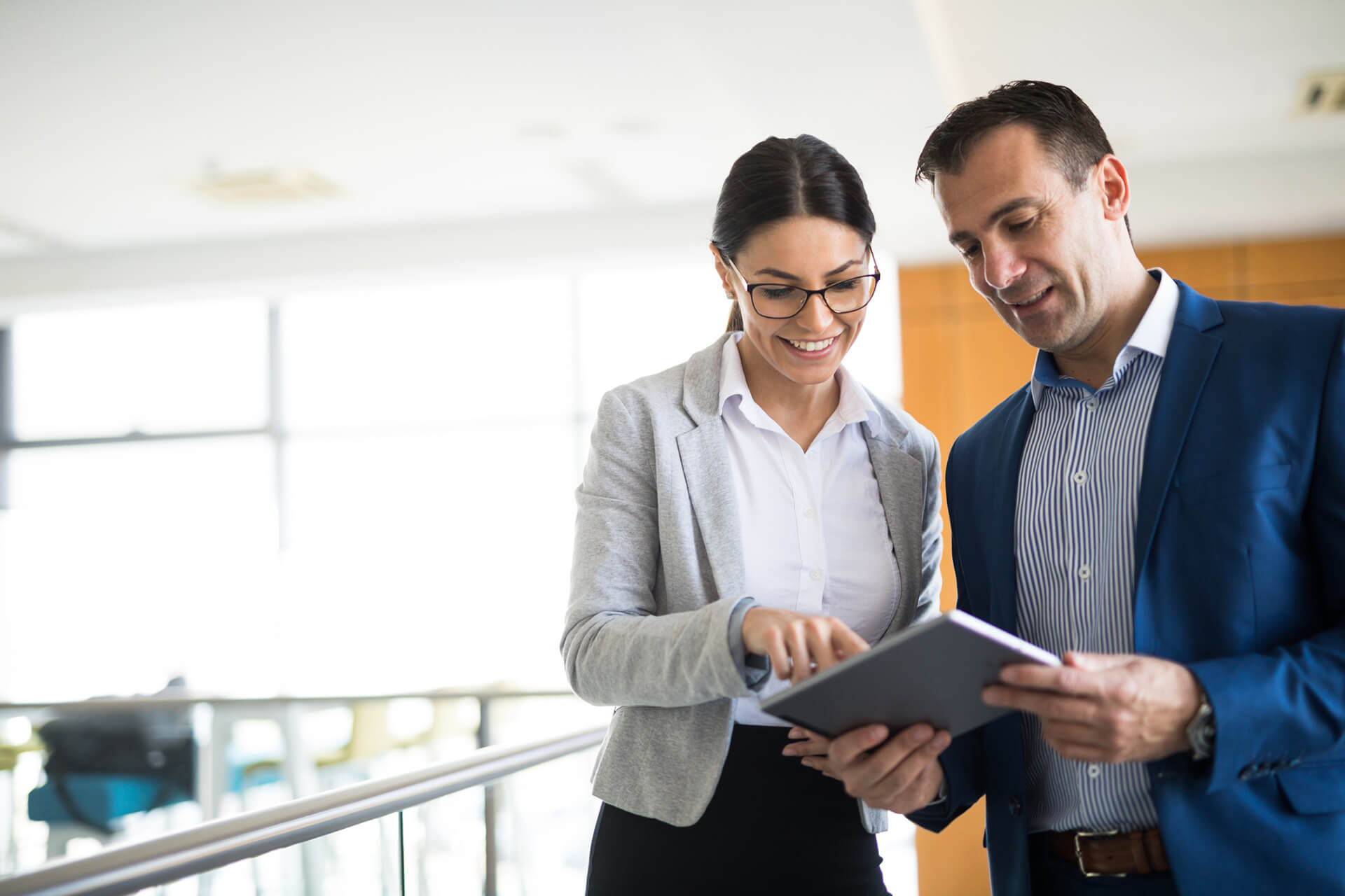 Smiling man and woman standing in a brightly lit office looking at a tablet device held in the man's hand.