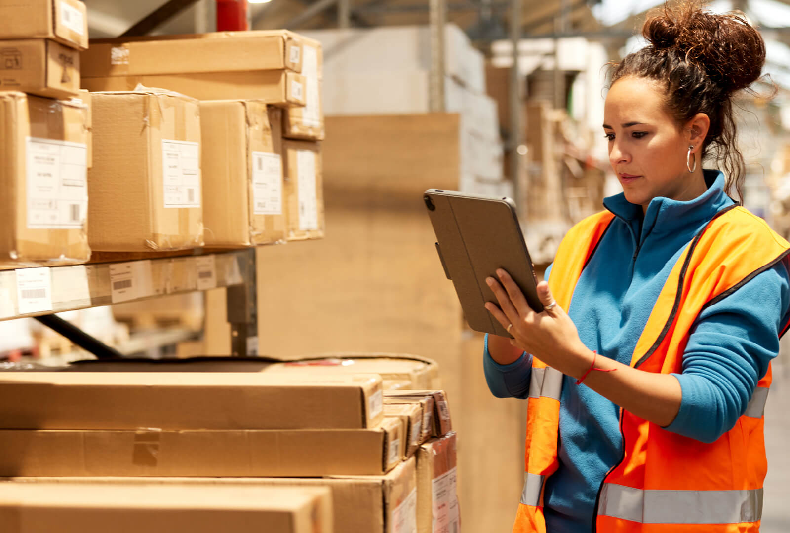 Woman in a warehouse wearing a orange reflective vest interacting with a tablet device.