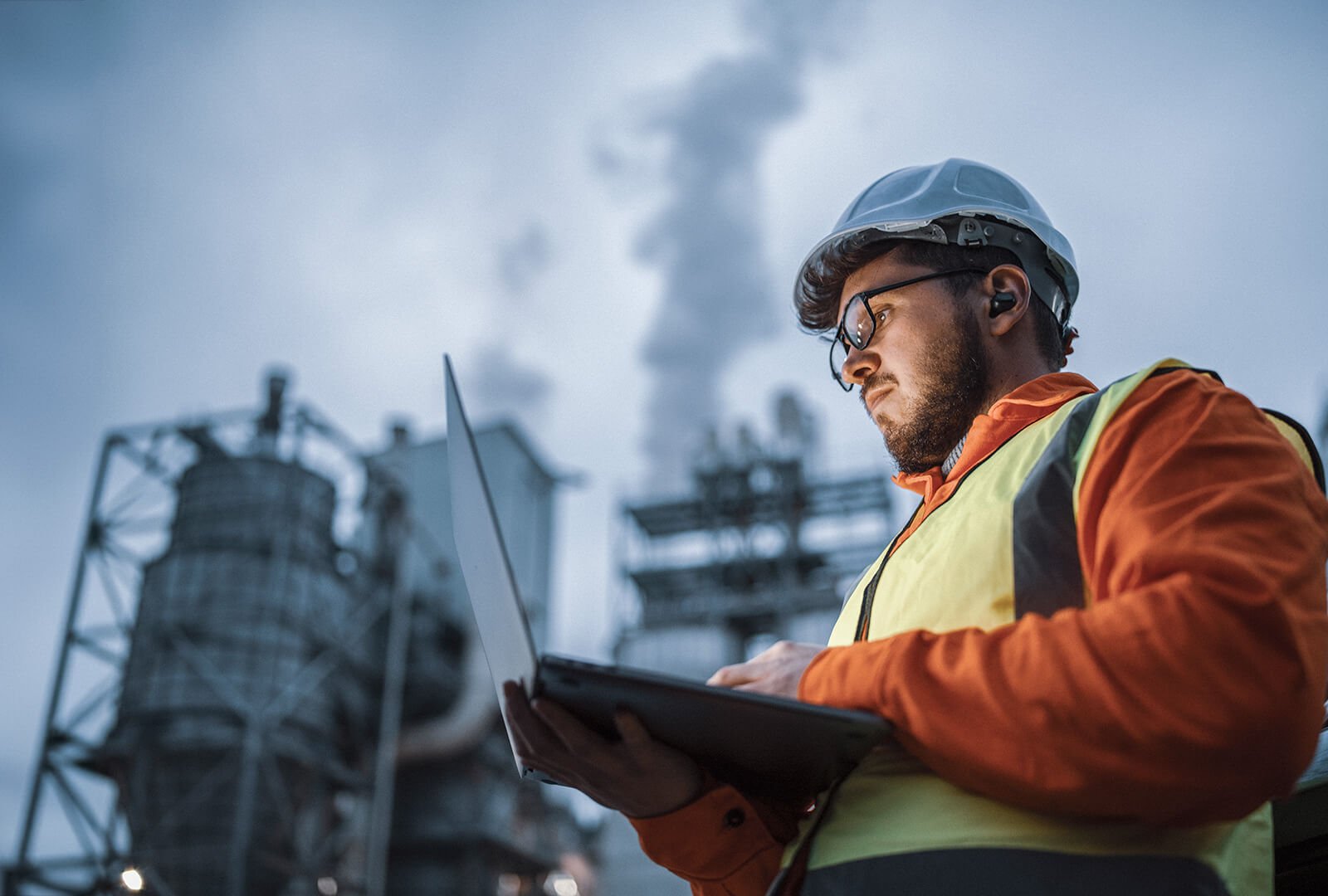 Man in a yellow safety vest and hardhat standing outside a factory working on a laptop held in his right hand.