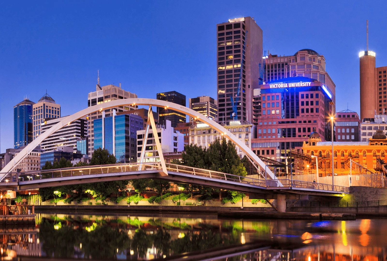 Cityscape of Melbourne behind a bridge with a neon blue Victoria University sign glowing atop a building.