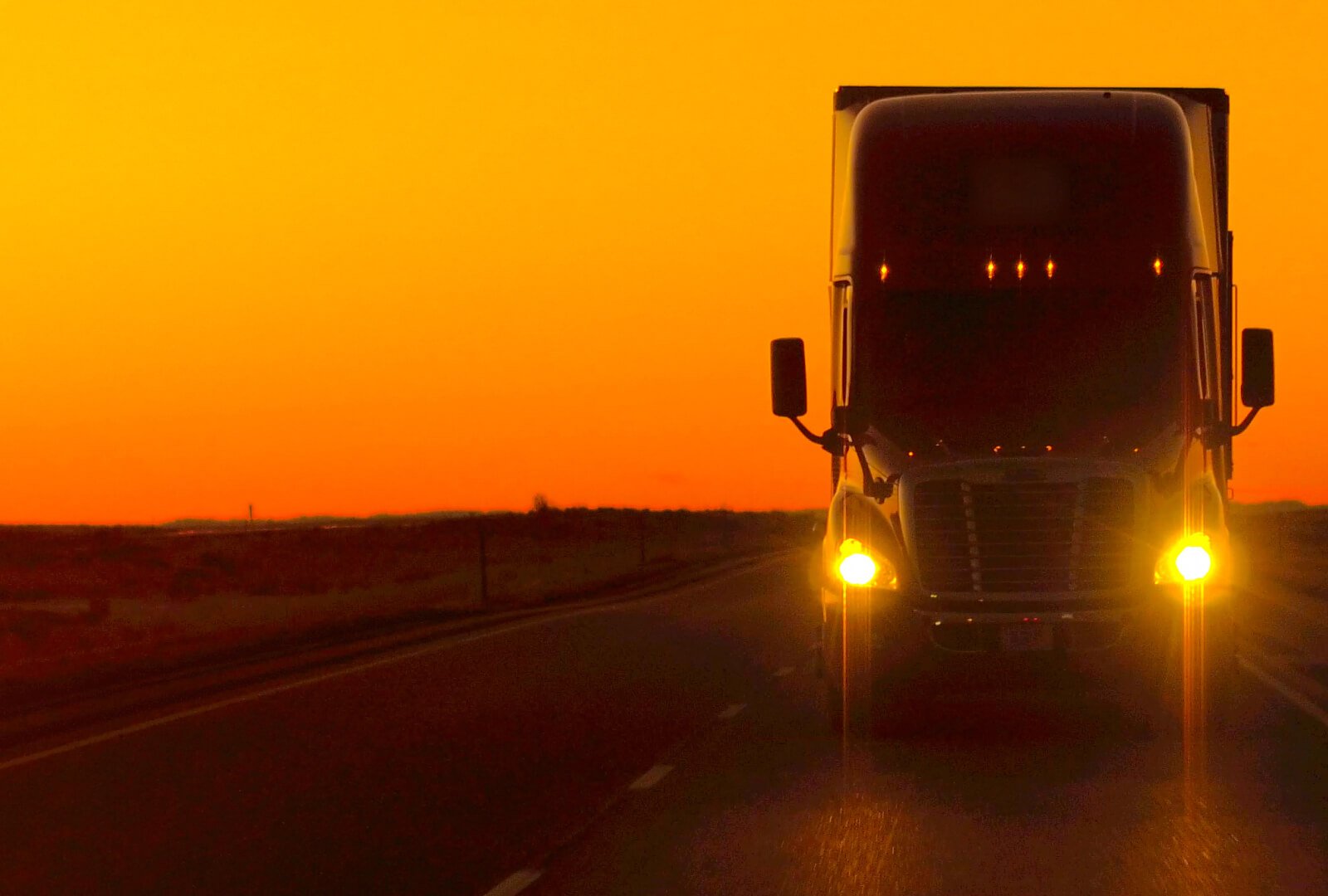 Photo of a tractor trailer truck motoring on a highway with an orange dusky sky in the background.