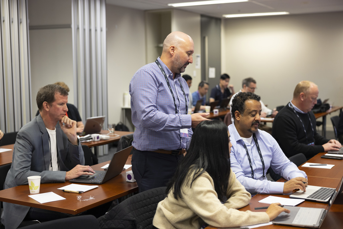 People sit at tables with laptops while a man stands in a conference room