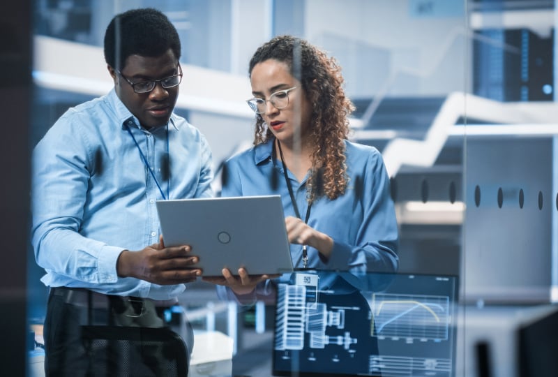 Two IT professionals reviewing data on a laptop in a modern technology operations center with digital dashboards and analytics displays visible in the background.
