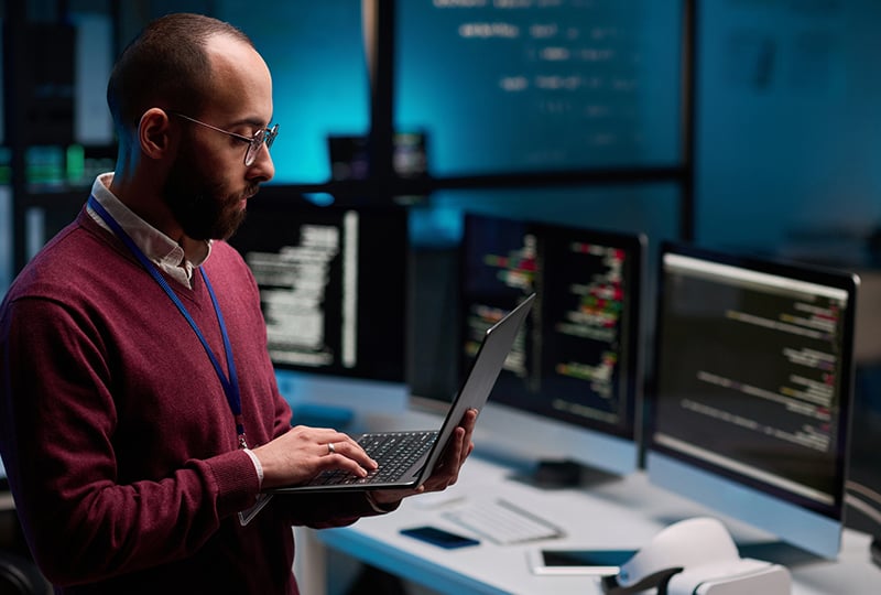 A man standing in front of multiple monitors displaying code in a dimly lit office. The individual is wearing a maroon sweater and holding a laptop, appearing to work or monitor computer systems in a technology or IT environment.