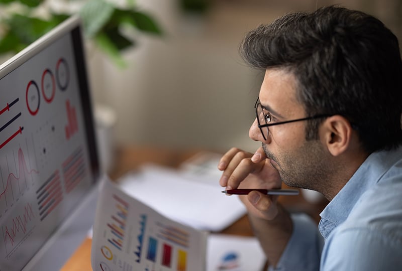 A man sitting at a desk, analyzing charts and graphs displayed on a computer monitor and printed on paper. The individual is holding a pen and appears to be deep in thought, working in a well-lit environment with plants in the background.
