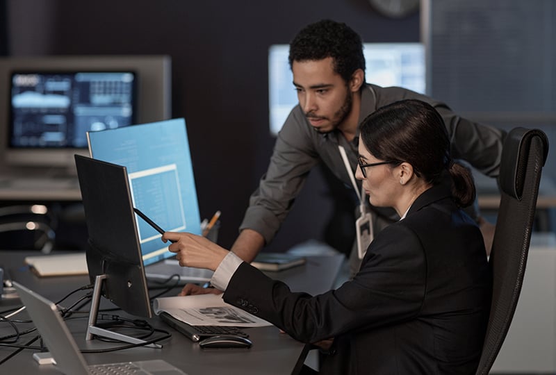 Two people working together in an office. One person is seated, pointing at a computer monitor with a pen, while the other is standing and leaning over the desk to look at the screen. Several monitors displaying data and documents are visible in the background, suggesting a collaborative work or analysis setting.