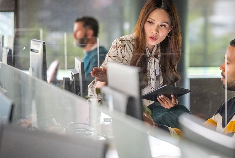 A woman holds a tablet beside a workstation with several monitors; two other people are visible behind glass partitions
