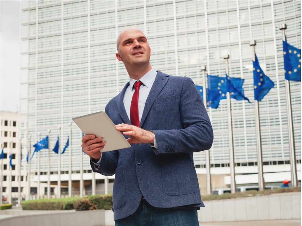 Person in a blue suit holding a tablet outside a modern building with multiple European Union flags in the background.