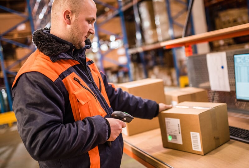 A man wearing an orange safety jacket scanning a box inside of a warehouse.