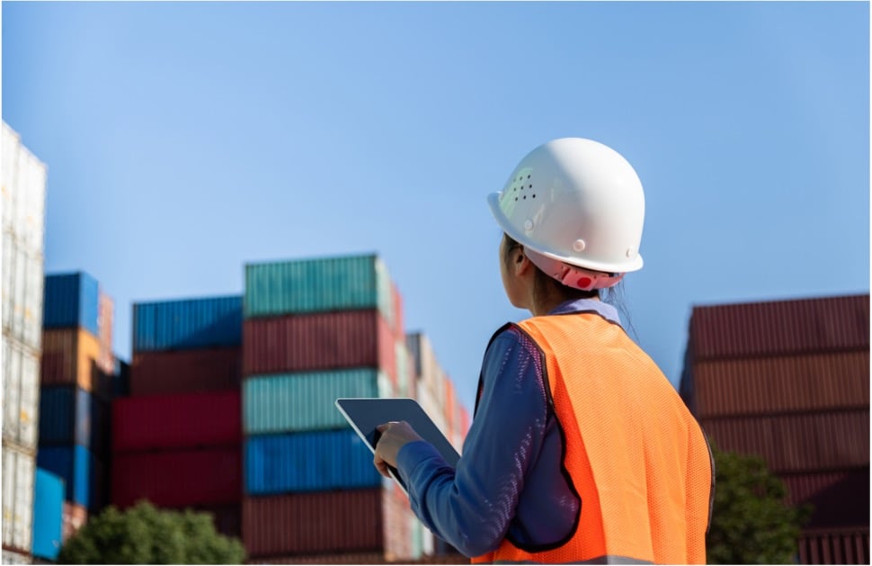 Worker wearing a hard hat and orange safety vest holding a tablet while inspecting stacked shipping containers in a cargo yard.