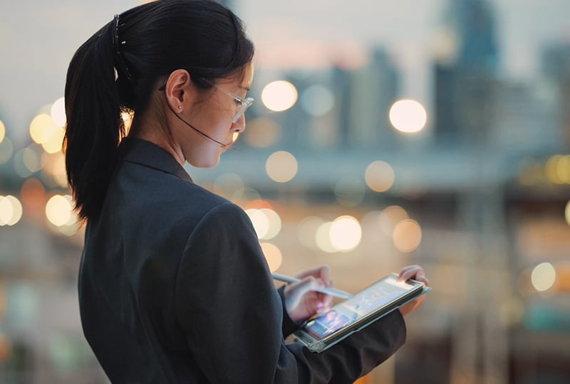 A woman dressed in a dark blazer, standing outdoors in an urban environment during twilight or evening. She is holding and using a tablet device, with blurred city lights and buildings in the background.