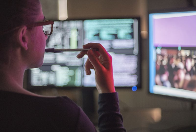 woman biting pen, thinking while looking at data on computers