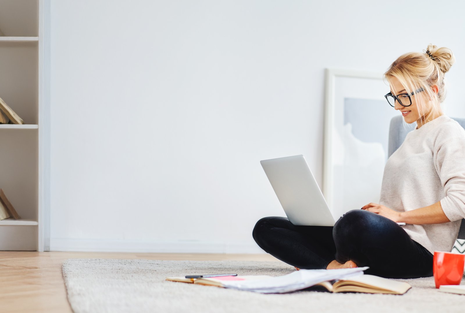 Woman sitting down on the floor in a white room working on a laptop.