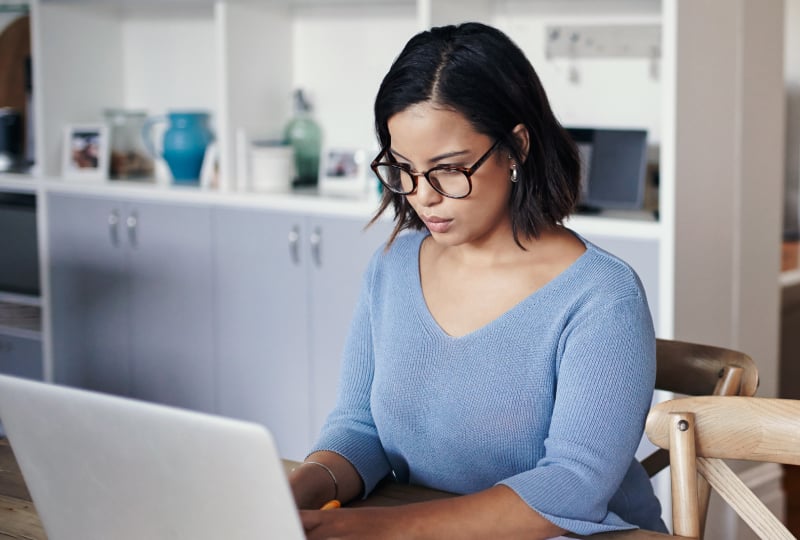 Woman sitting down at a table in a bright room working on a laptop.